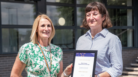 Two people standing outdoors in front of a building; one holds a framed certificate.